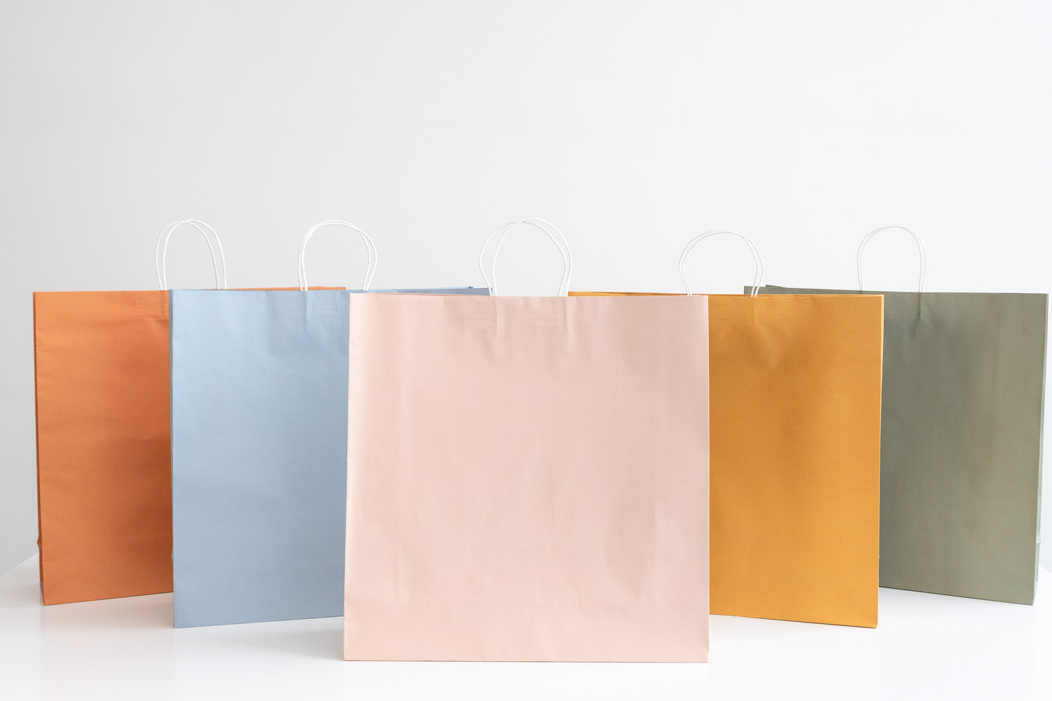 Earth Collection kraft carry bags in five vibrant colors: orange, blue, pink, yellow, and green. These recyclable and eco-friendly bags are perfect for sustainable packaging solutions.