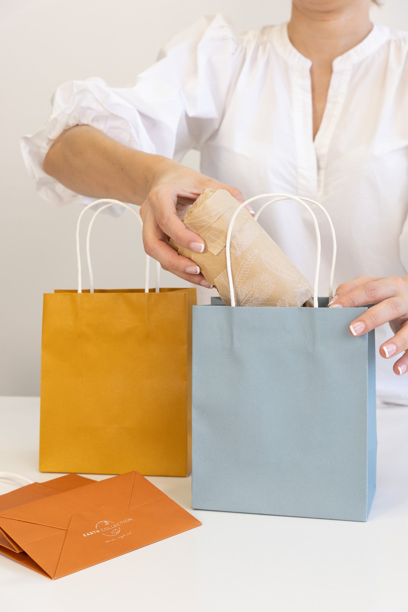 Three environmentally friendly kraft paper carry bags from the Earth Collection in light blue, burnt orange, and mustard yellow, showcasing sustainable packaging options.